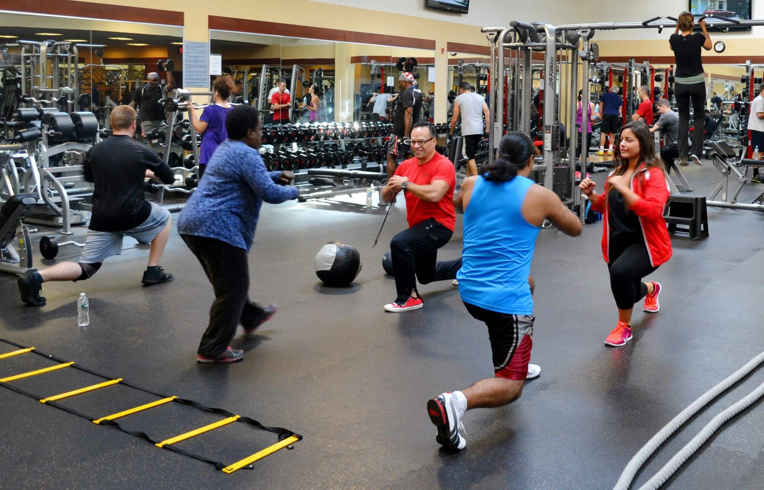 Small group fitness class participants engaging in various exercises, including lunges and medicine ball workouts, in a gym setting in Prairie Village, KS.